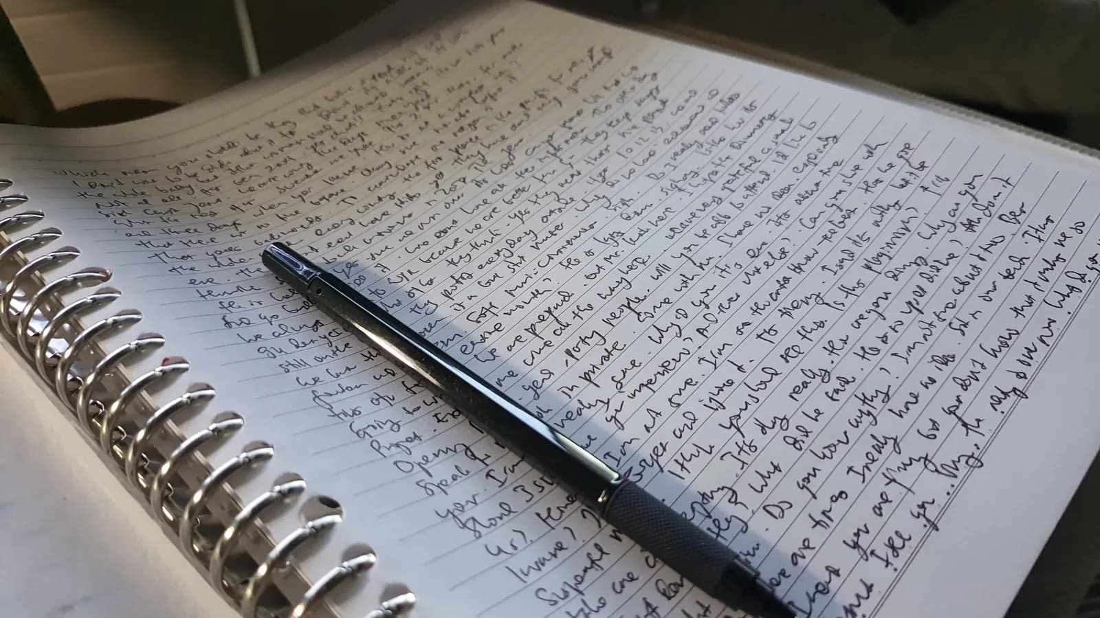 Hojas de papel escritas a mano junto a un cuaderno abierto y un bolígrafo sobre una mesa