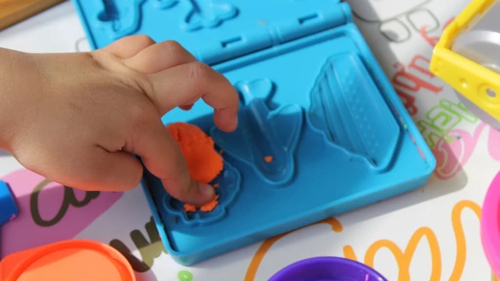Mano infantil apretando plastilina naranja en un molde azul, rodeada de material de manualidades
